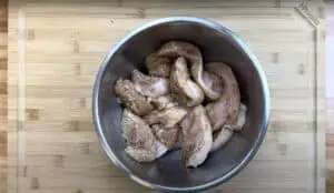 Marinated chicken strips in a stainless steel bowl on a wooden cutting board, ready for cooking.